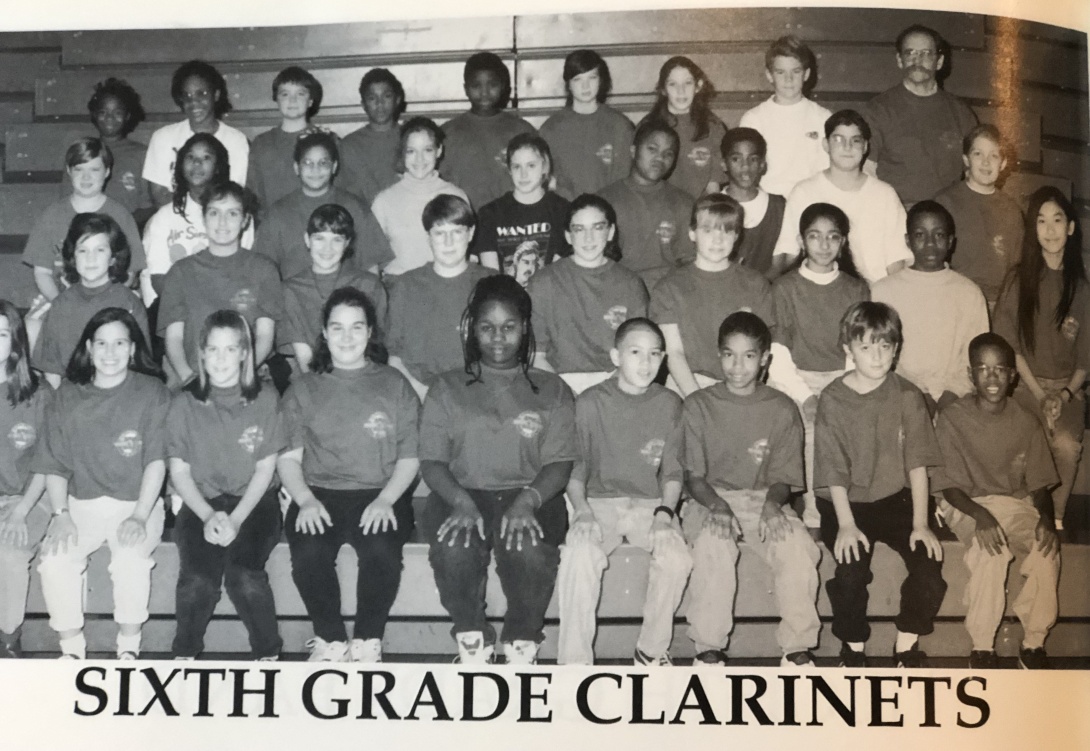 A black and white image showing four rows of 6th grade students facing the camera and sitting on bleachers. The only visible Indian American student is a girl sitting in the second from the bottom row. The text below the photo reads "Sixth Grade Clarinets." 
