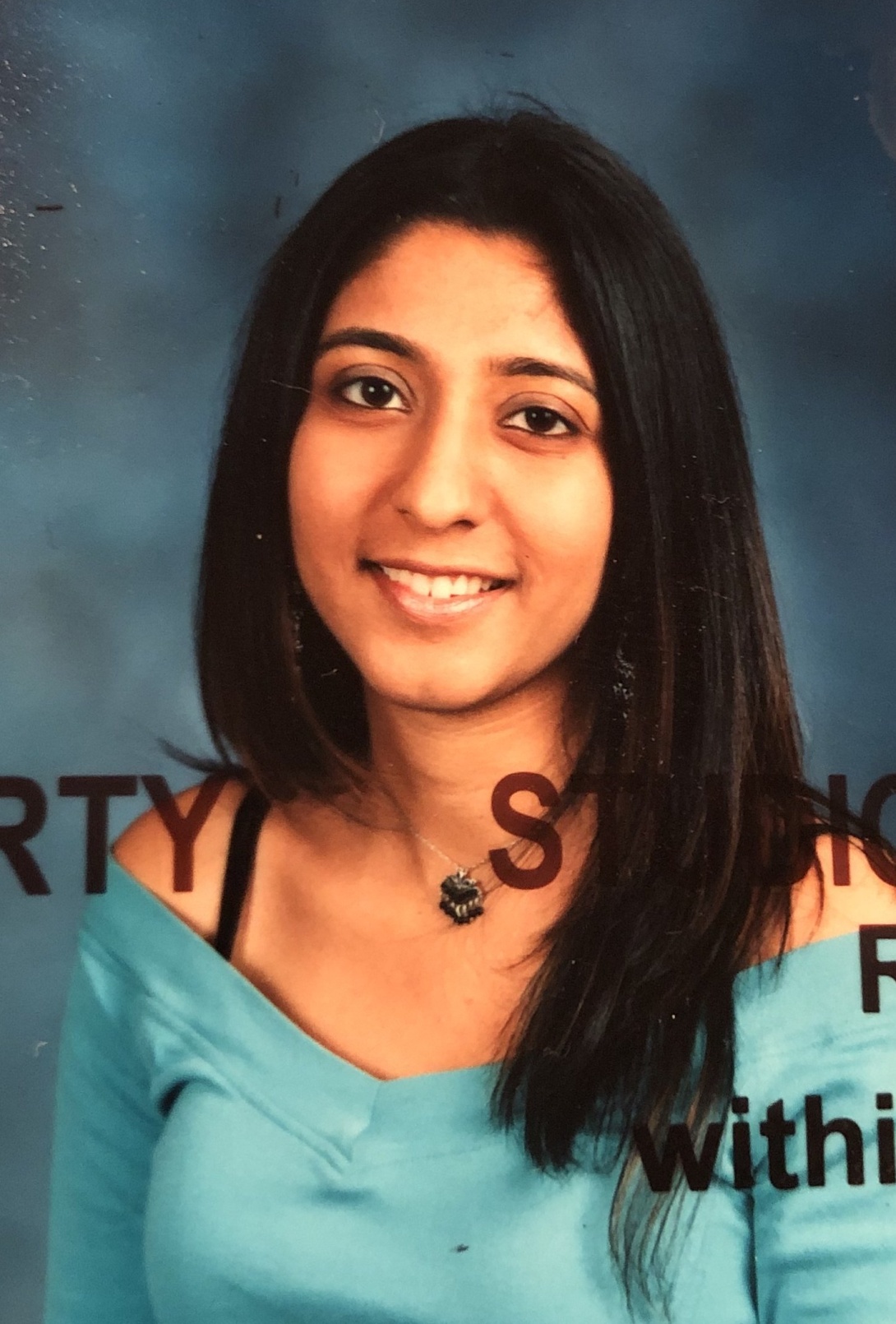 A young South Asian woman smiles at the camera for a studio portrait. She is wearing a blue, off-the-shoulder shirt.