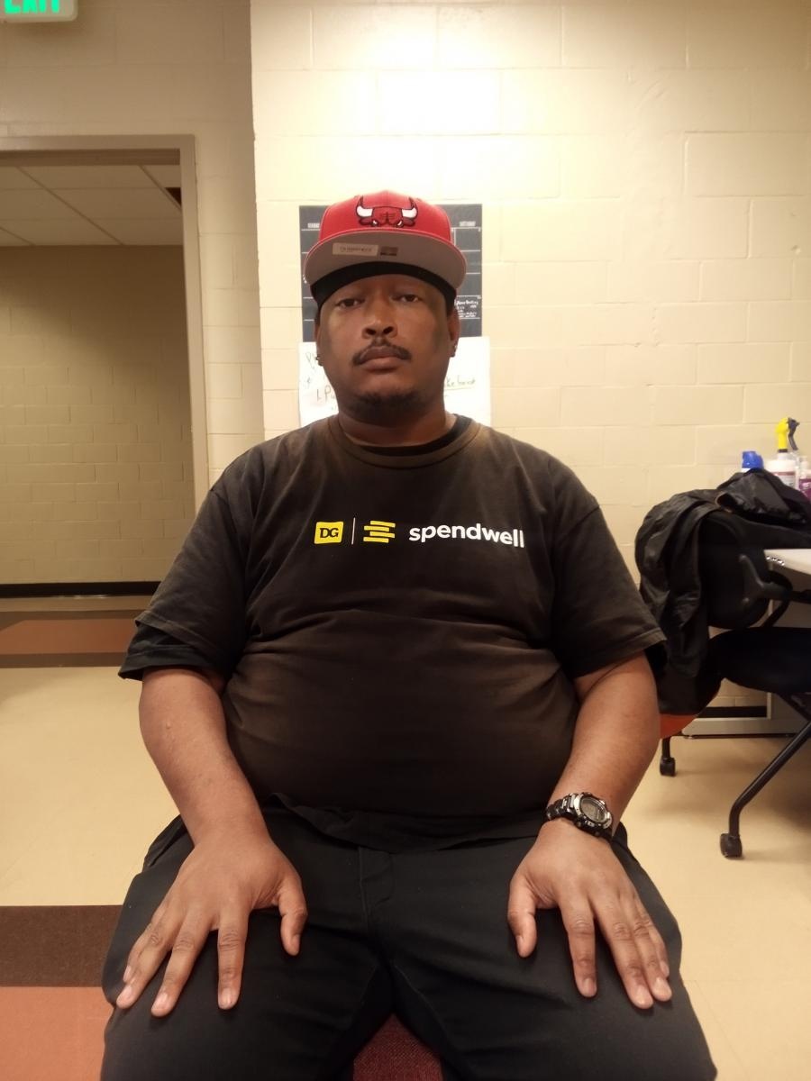 Black man in a ball cap and black clothing sits in a chair looking at the camera