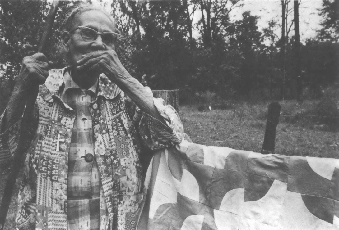 Black and white photo of elderly Black woman standing with walking stick next to hanging quilt, with her hand over her mouth in thought