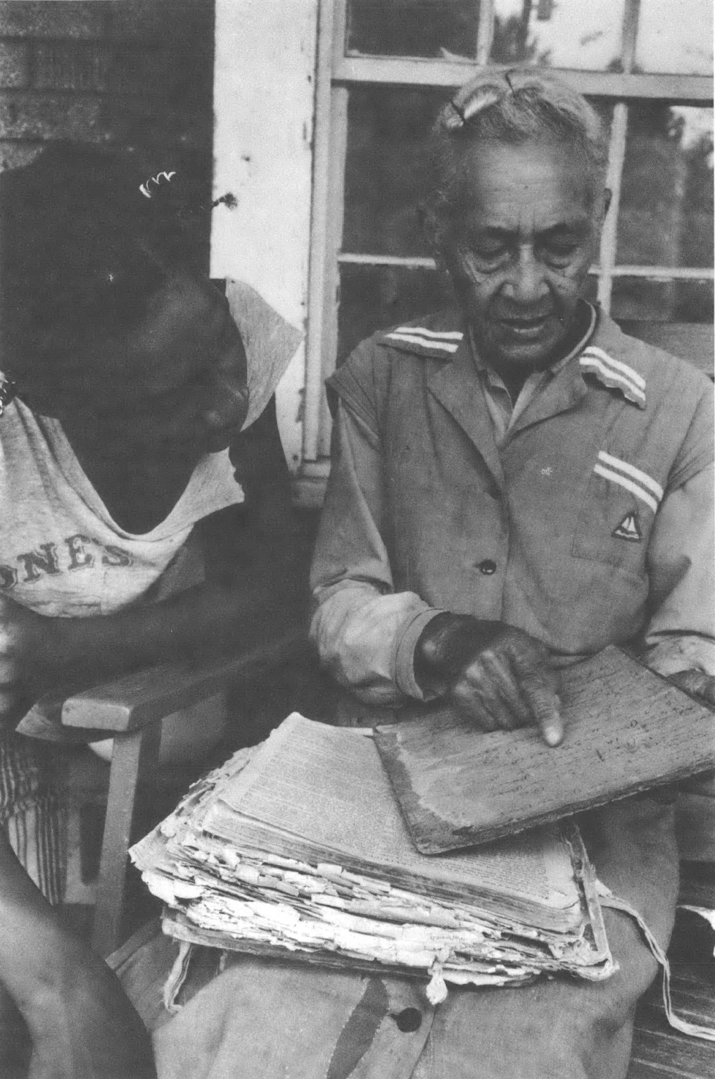 Black and white image of an older Black woman and her grandson sitting on the porch, the grandmother pointing at a document she is holding.