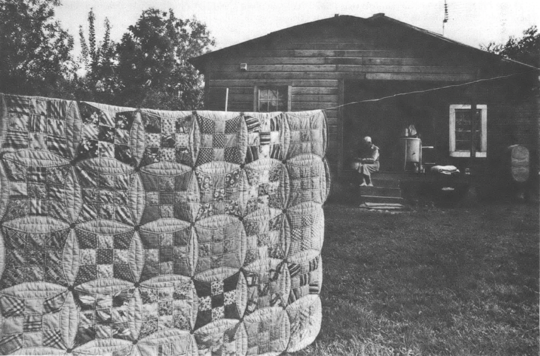 Black and white photo of quilt hanging outside of a house