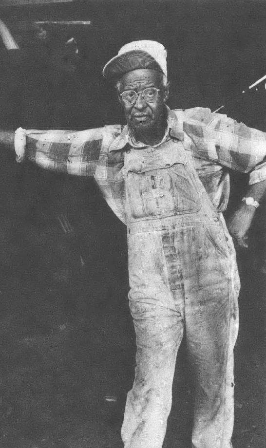 Black and white photo of Black man in cap, overalls, and plaid shirt leaning against a doorframe
