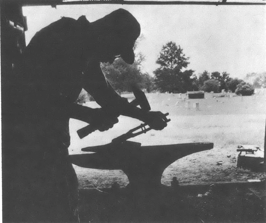 Black and white photo showing silhouette of a man hammering a piece of metal on an anvil