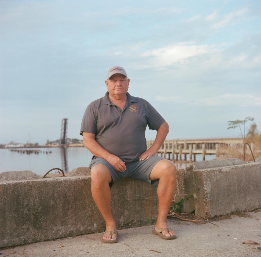 White man in ball cap, polo shirt, shorts, and flip flops sitting near lake's edge