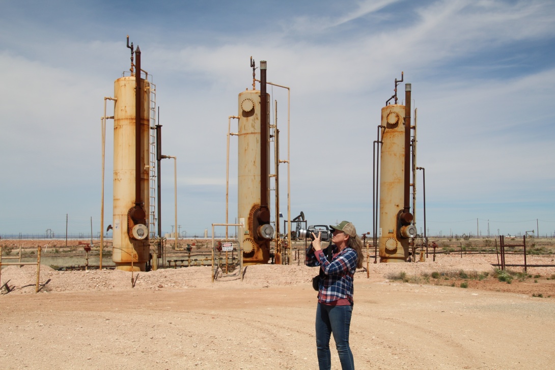 Woman in jeans and plaid shirt holding camera in front of her face, in front of three large oil wells in a desert-like landscape