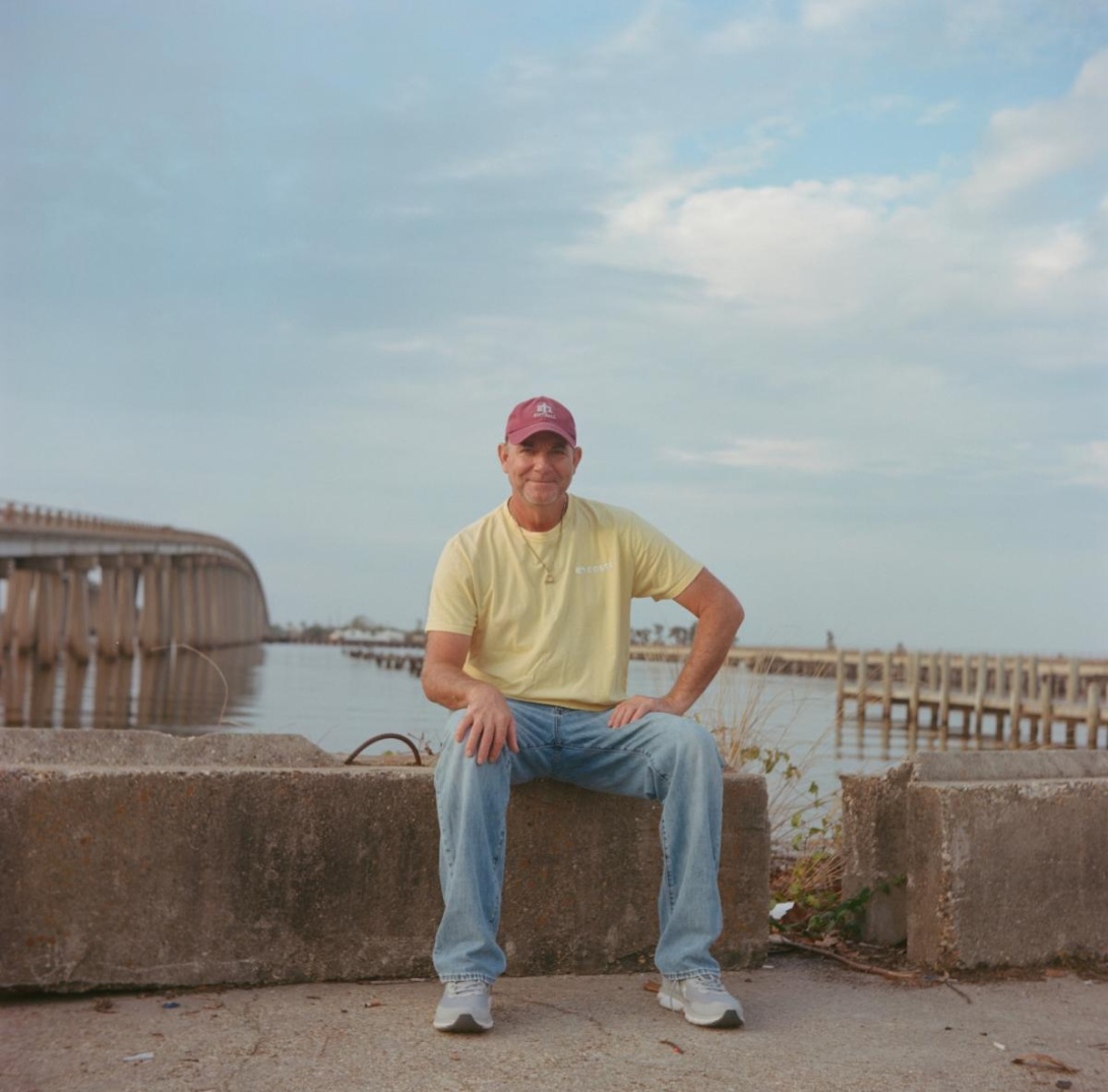 White man in ball cap, T-shirt, and jeans sitting near lake's edge