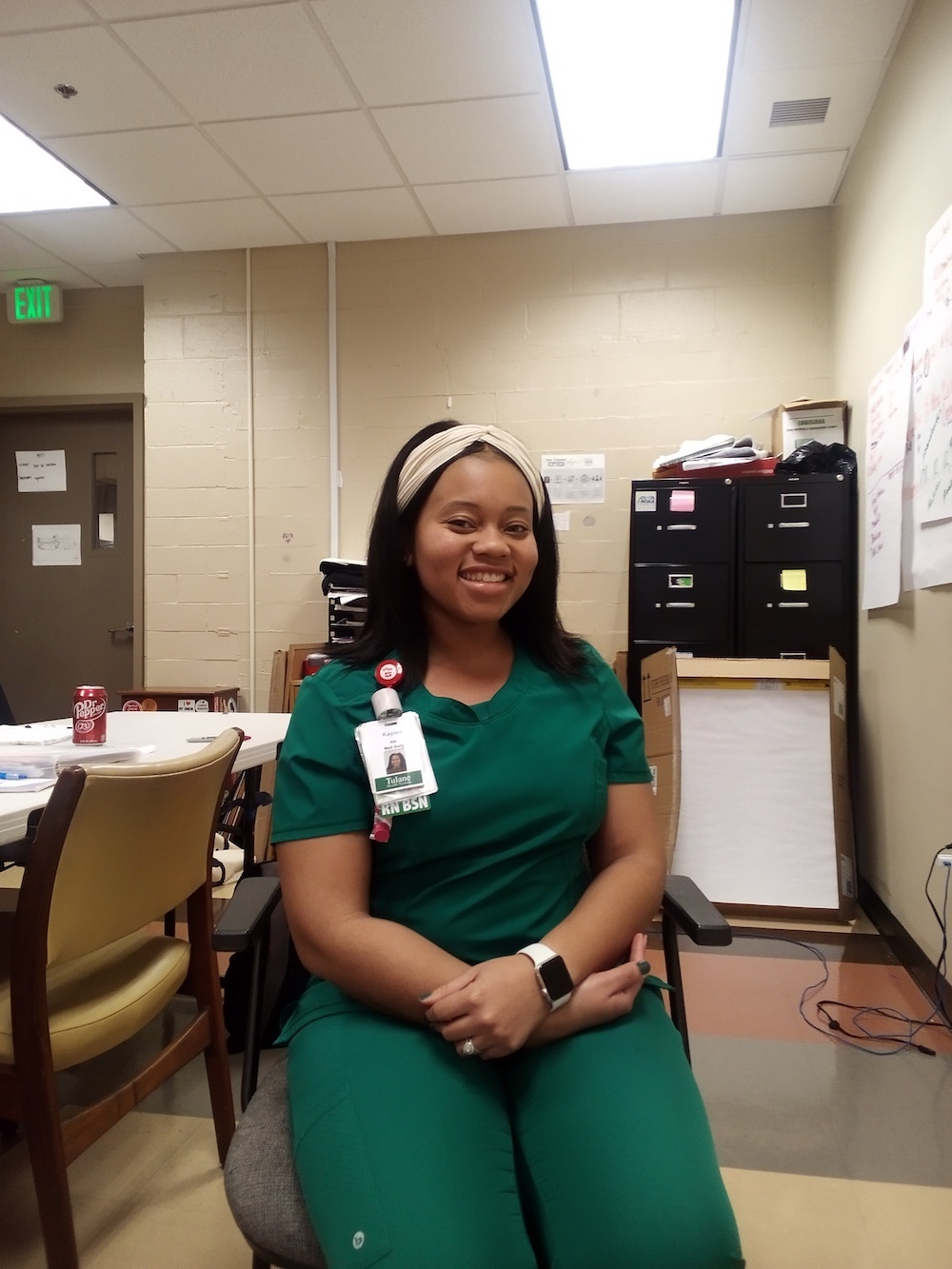 Black woman wearing green scrubs and a hair band, sitting in chair and smiling