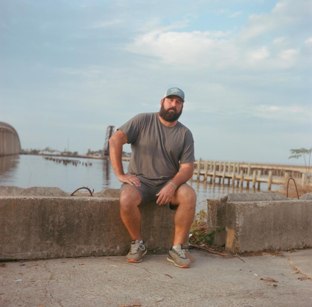 Bearded white man in T-shirt and shorts sitting near the lake's edge