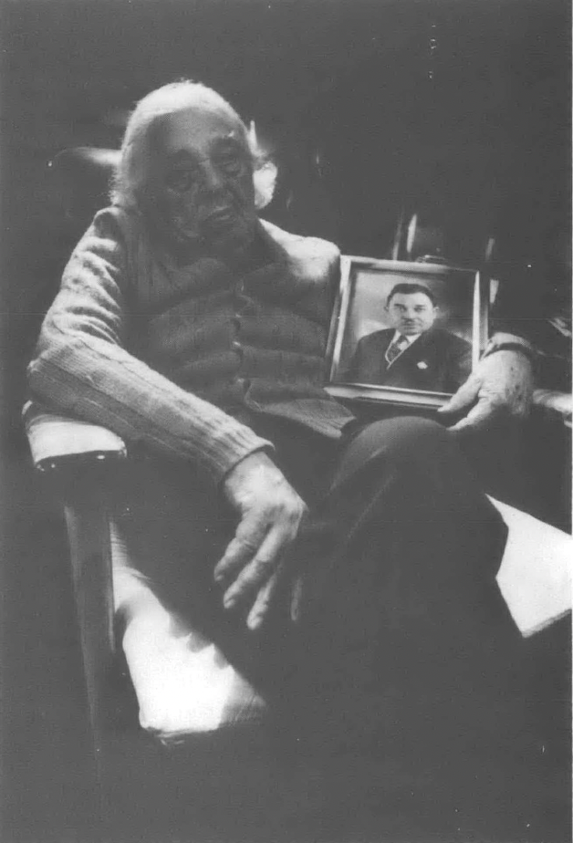 Black and white photo of old white man sitting in chair holding portrait of younger man