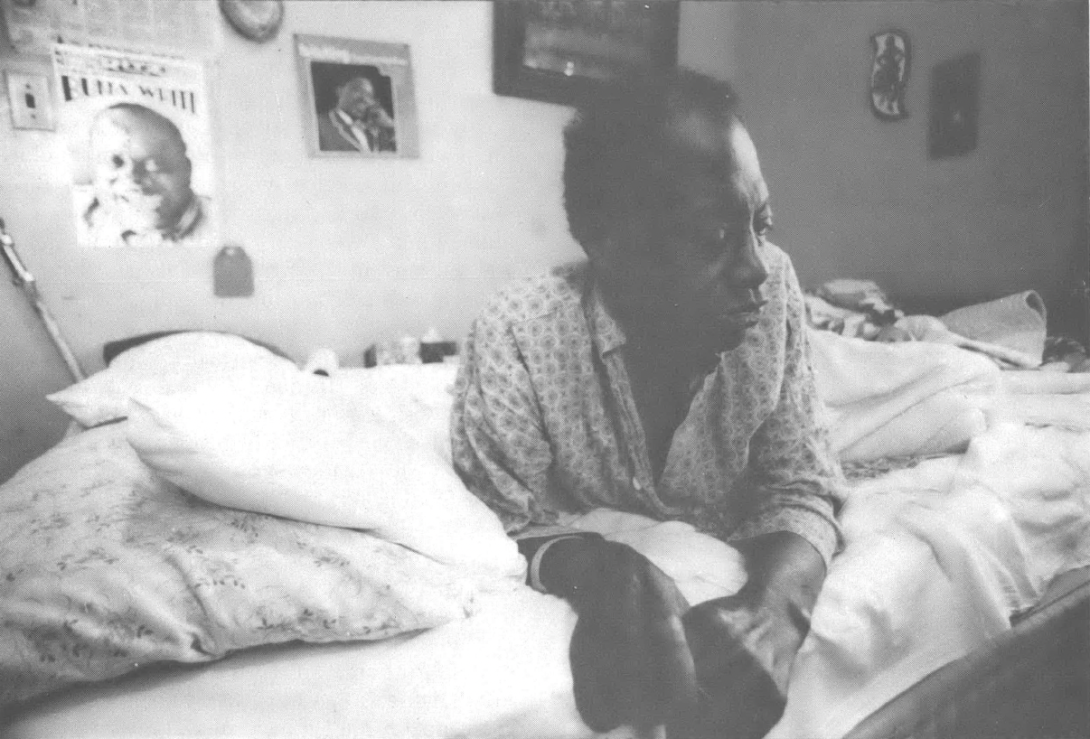 Black and white photo of Black man laying on his stomach on bed, looking down away from camera