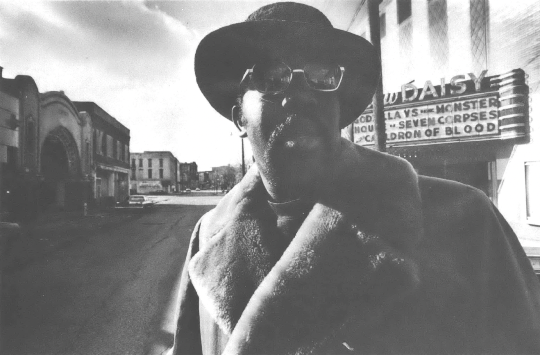 Black and white photo of Black man in hat and sunglasses and fur coat standing in the street