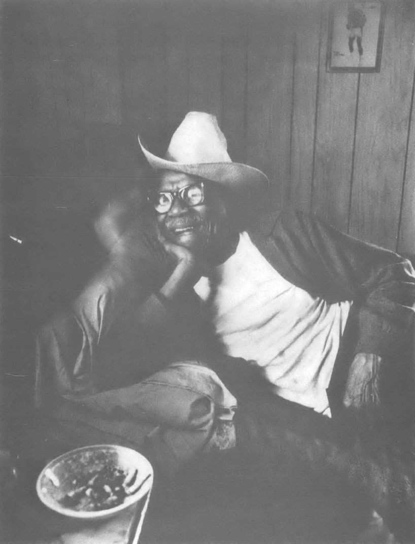 Black and white photo of Black man in cowboy hat and glasses reclining on a couch 