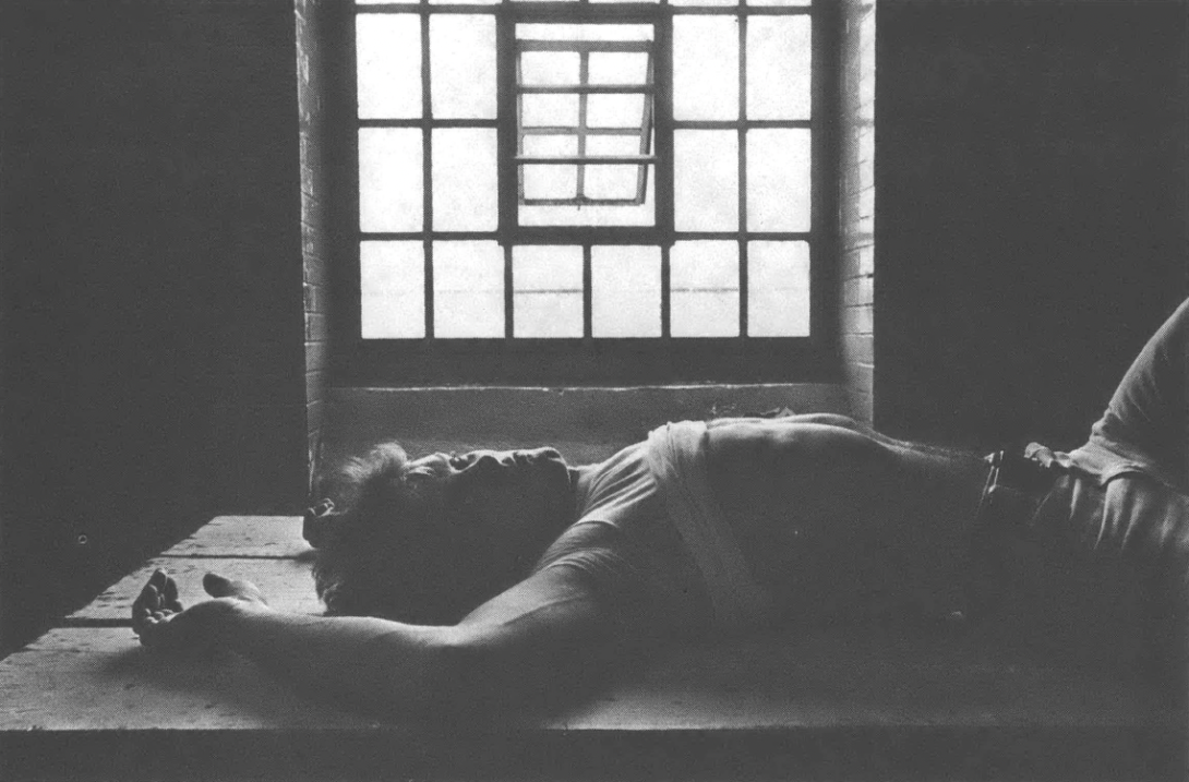 Black and white photo of man lying with arm above his head in front of a barred window