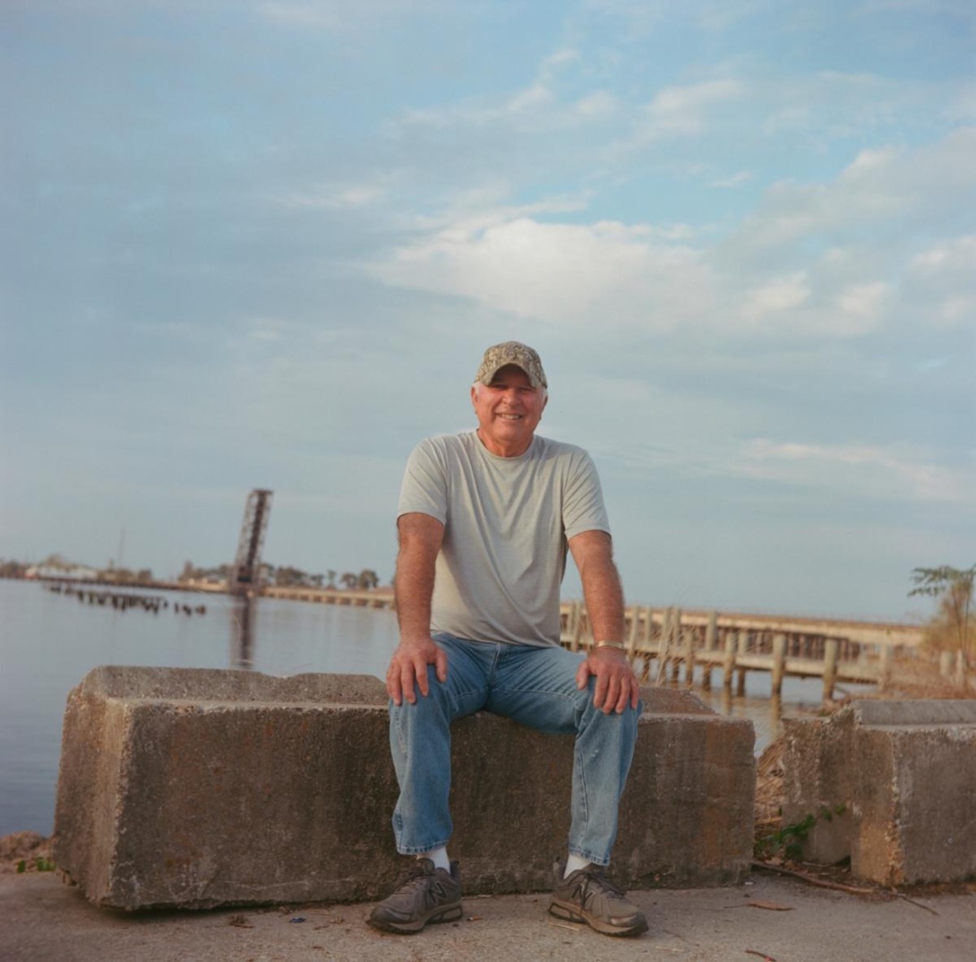 White-haired white man in ball cap, T-shirt, and jeans sitting near lake's edge