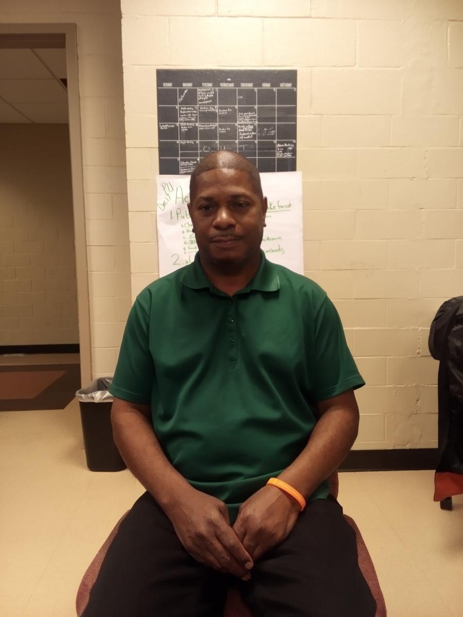 Black man in a green shirt and black pants sits in a chair looking at the camera