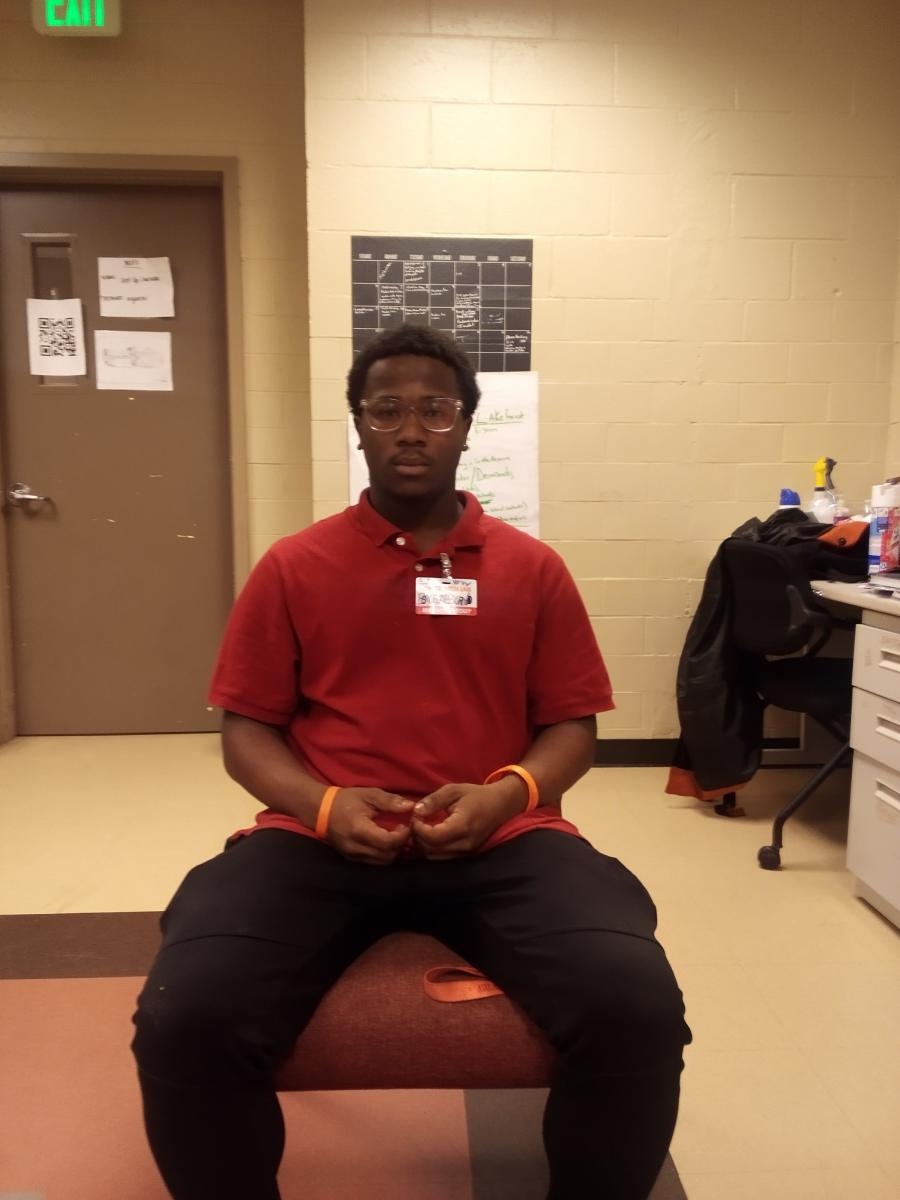 Young Black man wearing glasses, red shirt, black pants, sitting in chair looking at the camera