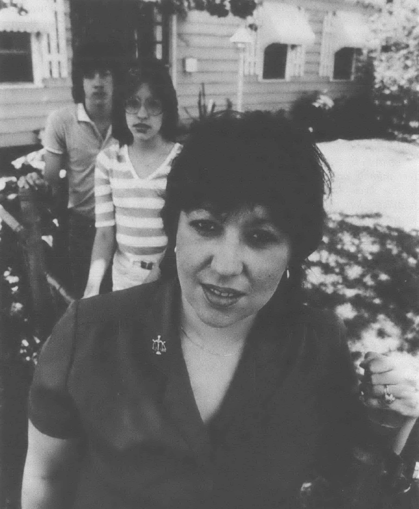 Black-and-white photograph of a woman with short hair, with two teenaged children standing behind her, looking directly at the camera and standing in front of a home.